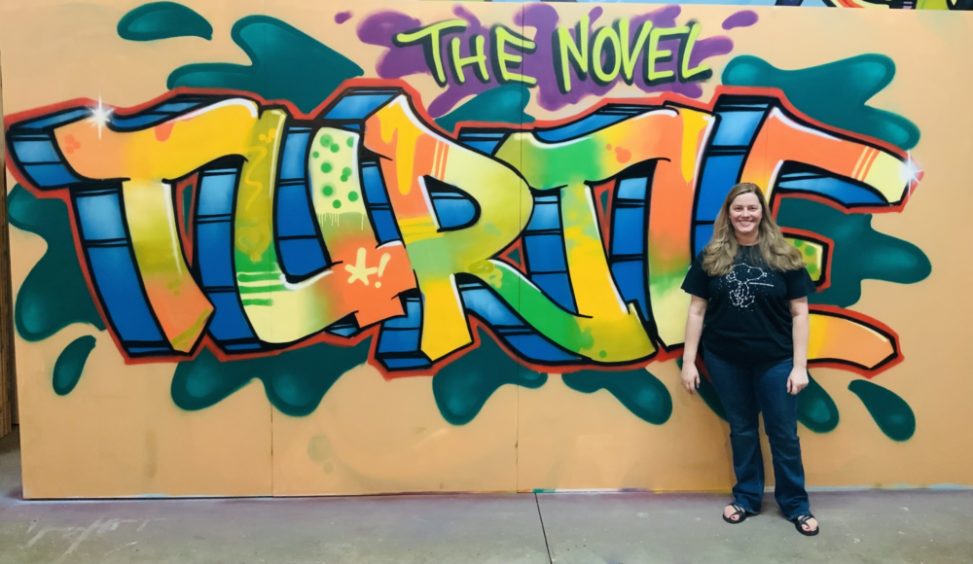 Lori Frye, a.k.a. The Novel Turtle, standing in front of a colorful graffiti painting that says "The Novel Turtle".