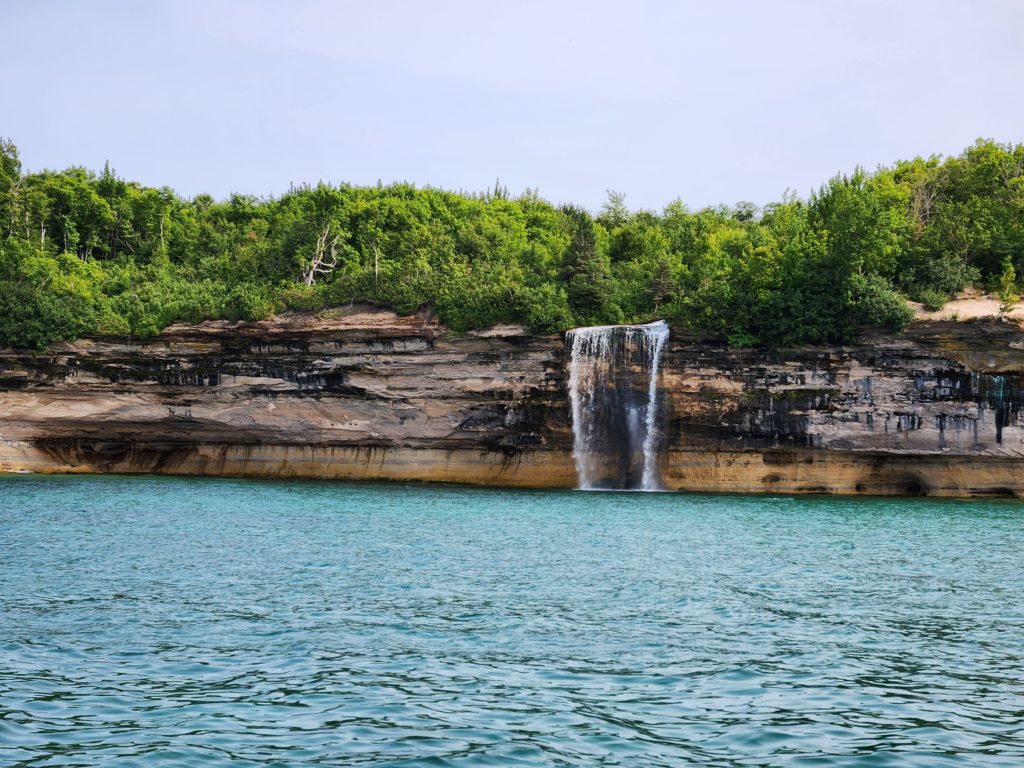 Spray Falls at the Pictured Rocks National Lakeshore. A waterfall that runs over the rock wall that makes up the lakeshore. Blue sky and a thick layer of trees are at the top of the picture, and Lake Superior lines the bottom of the picture. 