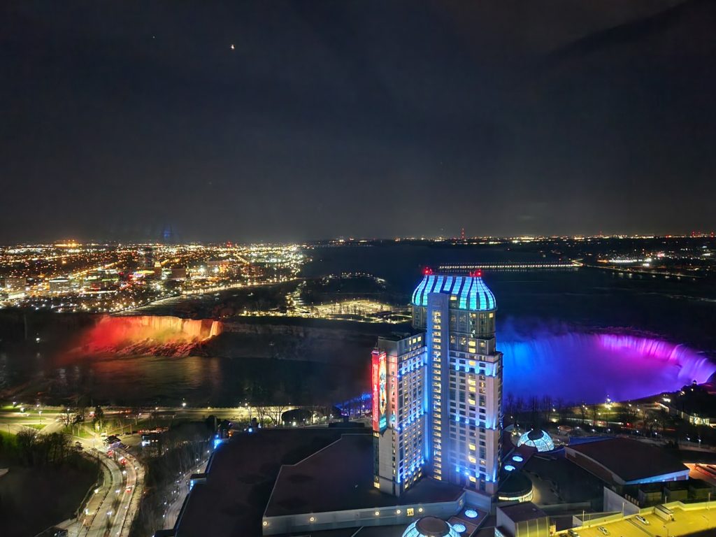 A view of the Niagara Falls lit up at night, with all of the city lights around it, from a hotel on the Canadian side. 