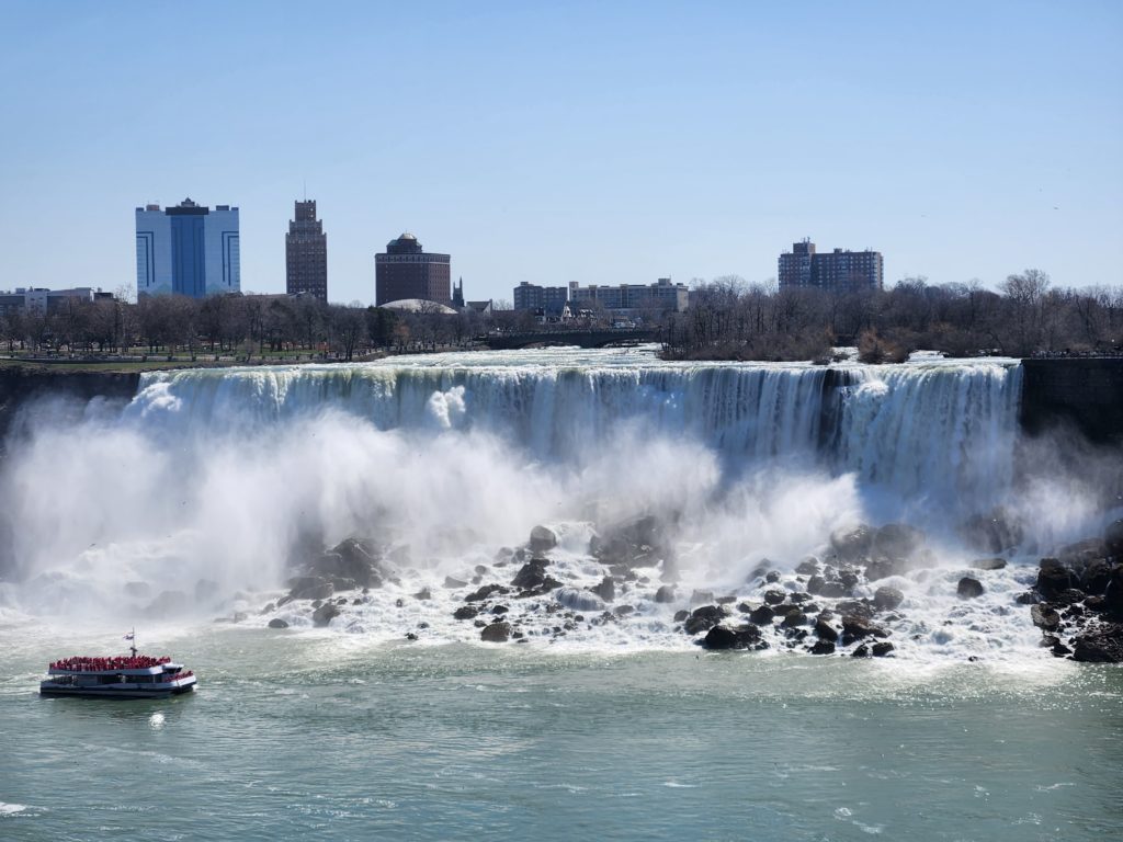 A view of the American side of Niagara Falls, with a tour boat going by it. Tall hotels off in the distance. 