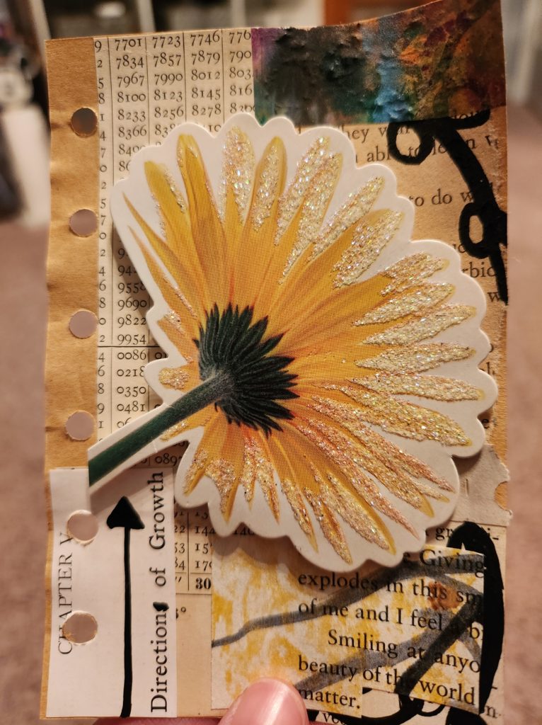 A 3x5 piece of paper with a mixed media collage on it. Different vintage papers make up the background, while a large gerbera daisy stands out in the middle. 