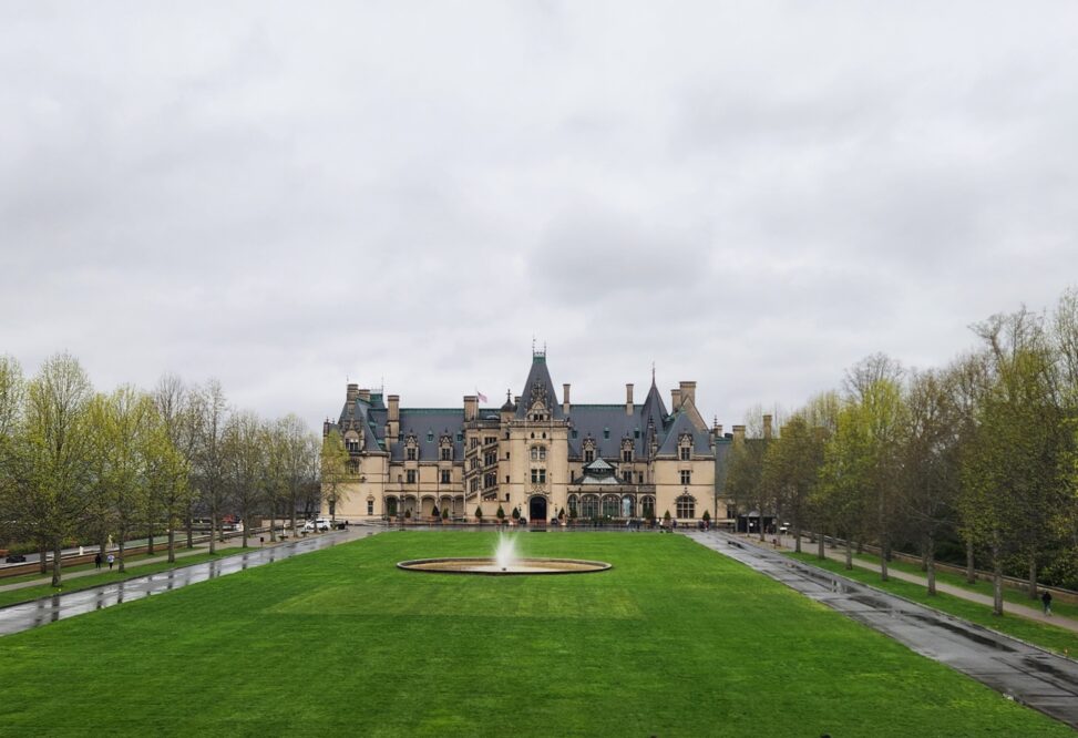 Looking at the Biltmore House from across the long front yard that runs in front of it. Sidewalks and a paved road run along both sides of the grass and there is a fountain in the center of the lawn.