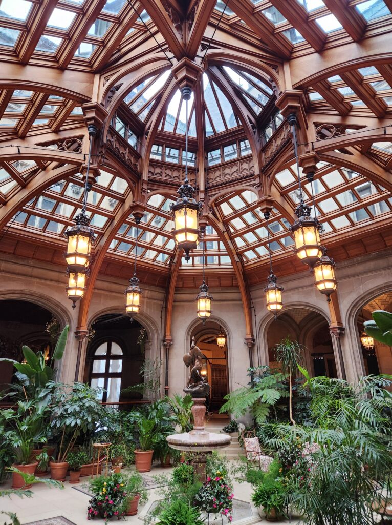 The Winter Garden in the Biltmore Estate. A circular room with arched doors all around the "walls". The ceiling is an intricate pattern of clear glass that allows the sunlight to shine through. The room is filled with large plants and has a fountain in the middle of it.