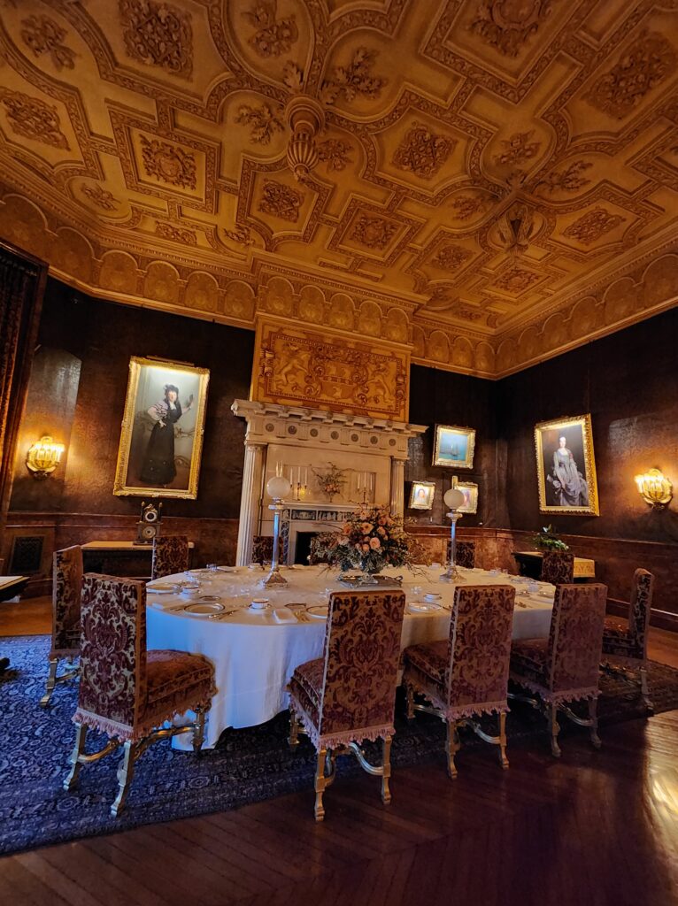 A dining table with eleven chairs around it. The room around it has a fireplace and is ornately decorated. 