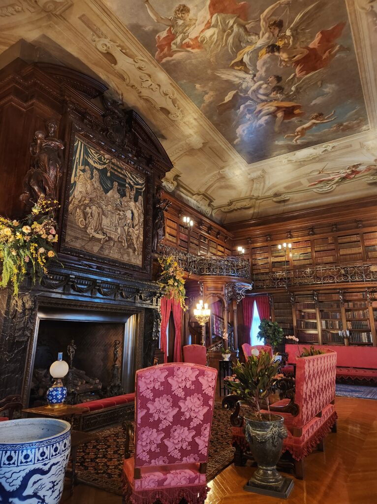 The library at the Biltmore Estate. A room filled with bookshelves all around, a large fireplace on the left (with a painting above it), chairs and tables here and there for reading and studying, and a ceiling with an elaborate painting. 