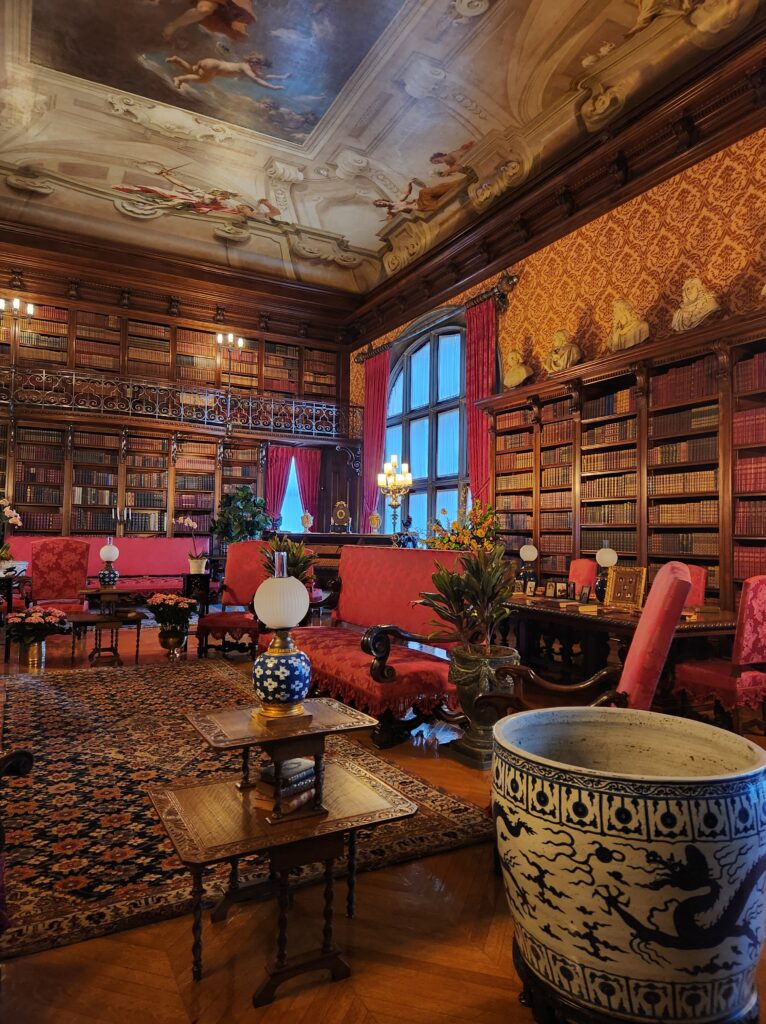 The library at the Biltmore Estate. The room is filled with bookshelves that are filled to the brim with books. A balcony runs along three walls of the room (going above the fireplace in one spot). There are chairs and tables all around the room. 