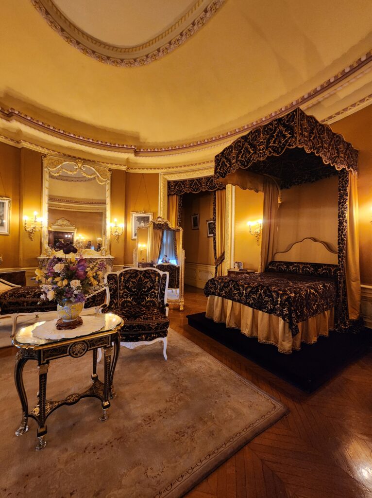 Edith Vanderbilt's bedroom at the Biltmore Estate in Asheville, NC. A canopy bed sits to the right. A large dressing mirror stands near it. There is a fireplace with a large mirror over top of it. a fainting couch, chair, and small table sit near the center of the room. The entire room is decorated in gold, purple and has wooden floors. 