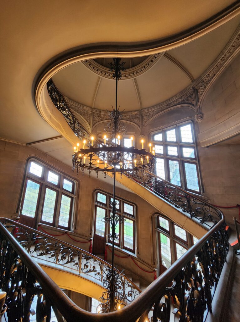 One of the grand spiral staircases that go between the floors of the Biltmore House. There is a large chandelier that hangs from the top floor ceiling, all the way through the spiral of the staircase down to the bottom floor. 