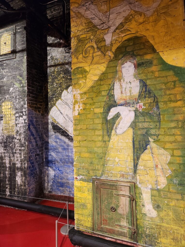 A painted wall in the Halloween Room of the Biltmore Estate that depicts a woman standing with her hands in front of her. She's wearing a simple dress with a cape style jacket over top of it. 