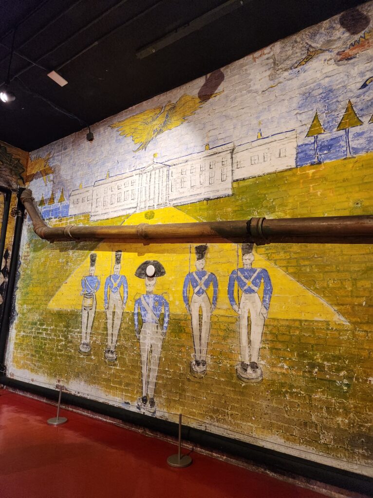 A painting on the wall of the Halloween Room of the Biltmore Estate that depicts five soldiers standing in formation in front of a large building. One of the soldiers is playing a drum. 