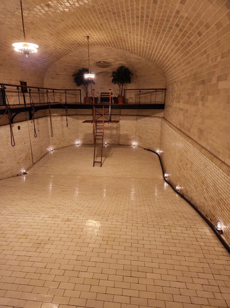 The swimming pool that is in the basement of the Biltmore House. Now drained of water because of leaks, it still has the original lighting around the bottom, diving board, and railing around the side. The pool is made out of white bricks and is in an enclosed room with an arched ceiling. 